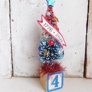 May include: A miniature bottle brush tree featuring a red top and blue bottom, decorated with small stars, beads, and a banner reading 1776 * 2026. The tree is topped with a blue star and sits on a wooden block featuring the blue number 4.