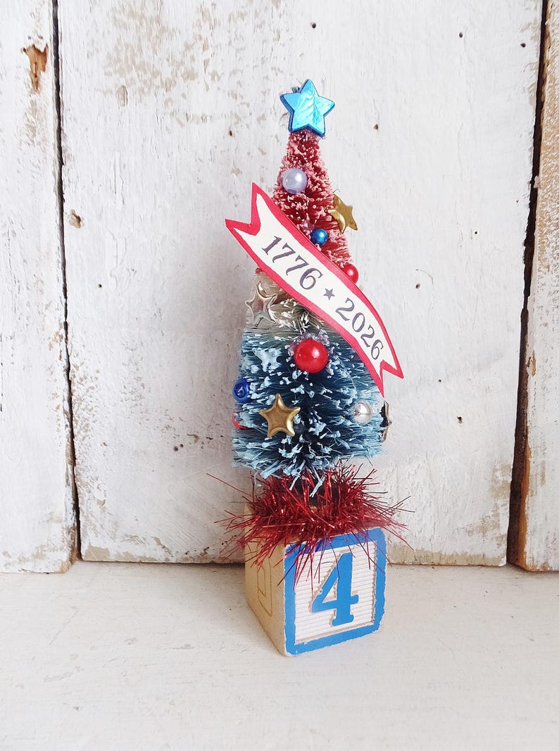 May include: A miniature bottle brush tree featuring a red top and blue bottom, decorated with small stars, beads, and a banner reading 1776 * 2026. The tree is topped with a blue star and sits on a wooden block featuring the blue number 4.