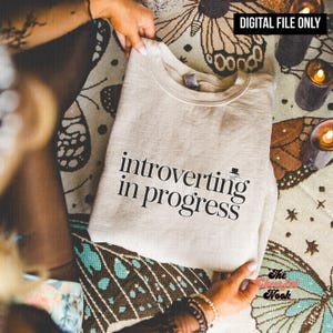 May include: A beige sweatshirt with the text "introverting in progress" in black. The sweatshirt is folded and held by hands. The words "DIGITAL FILE ONLY" are in a black rectangle in the top right corner.