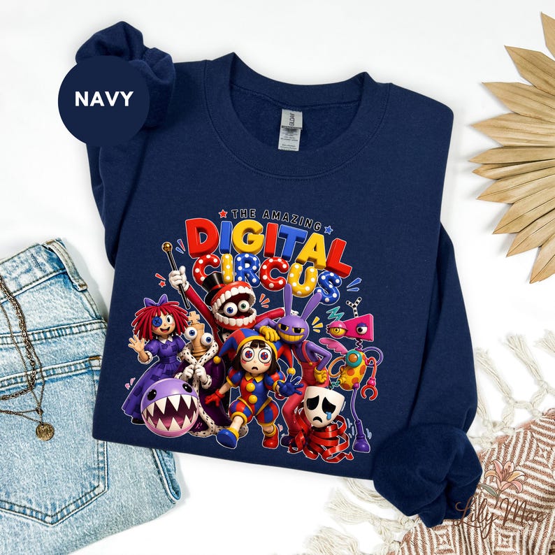 K&ouml;nnte beinhalten: Marineblaues Sweatshirt mit einer farbenfrohen Grafik von "The Amazing Digital Circus". Das Design enth&auml;lt den Text "THE AMAZING DIGITAL CIRCUS" in Rot, Blau und Gelb. Das Sweatshirt hat einen Rundhalsausschnitt und lange &Auml;rmel.