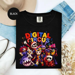 K&ouml;nnte beinhalten: Schwarzes T-Shirt mit einem farbenfrohen Grafikdesign von Cartoon-Charakteren und dem Text "DIGITAL CIRCUS" in Rot, Gelb und Blau. Das T-Shirt hat einen Rundhalsausschnitt und ein "Comfort Colors"-Etikett.