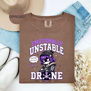 May include: Espresso-colored t-shirt with the words "Emotionally Unstable" and "Drone" in a purple and white design. The graphic features a cartoon character with purple hair and a black beanie. The shirt is surrounded by a hat, jeans, and boots.