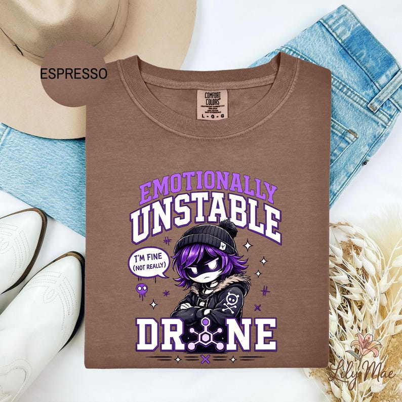 May include: Espresso-colored t-shirt with the words "Emotionally Unstable" and "Drone" in a purple and white design. The graphic features a cartoon character with purple hair and a black beanie. The shirt is surrounded by a hat, jeans, and boots.