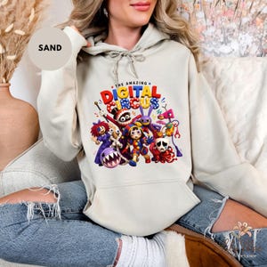 K&ouml;nnte beinhalten: Sandfarbener Kapuzenpullover mit einer farbenfrohen Grafik von Charakteren aus "The Amazing Digital Circus". Das Design enth&auml;lt den Text "THE AMAZING DIGITAL CIRCUS" in Rot, Blau und Gelb. Der Hoodie hat eine Kapuze mit Kordelzug und eine Vordertasche.