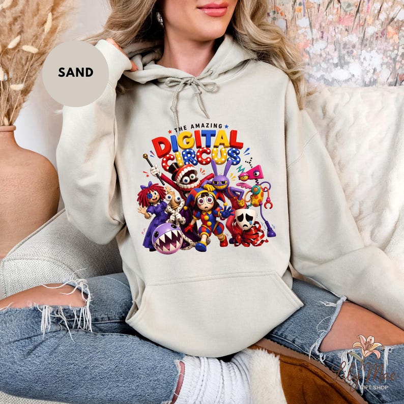 K&ouml;nnte beinhalten: Sandfarbener Kapuzenpullover mit einer farbenfrohen Grafik von Charakteren aus "The Amazing Digital Circus". Das Design enth&auml;lt den Text "THE AMAZING DIGITAL CIRCUS" in Rot, Blau und Gelb. Der Hoodie hat eine Kapuze mit Kordelzug und eine Vordertasche.