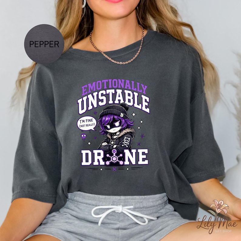 May include: Dark gray sweatshirt with the text "Emotionally Unstable Drone" and a cartoon character with purple hair and a black beanie. The character has a skull and crossbones on their jacket. The words "I'm fine (not really)" are in a speech bubble.