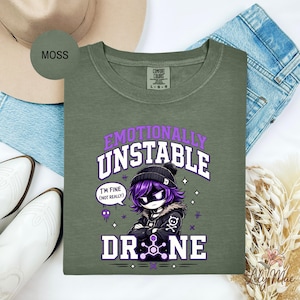 May include: Moss green long-sleeve shirt with the text "Emotionally Unstable Drone" in purple and white. The graphic features a cartoon character with purple hair and a black beanie. The shirt is laid on top of blue jeans.