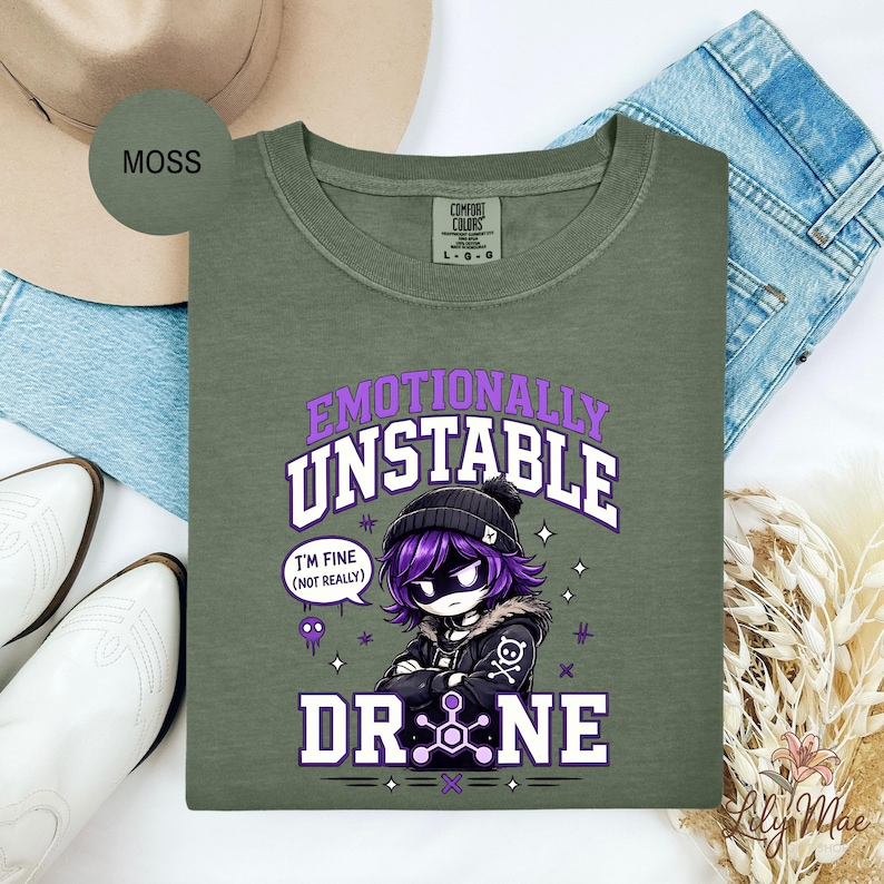 May include: Moss green long-sleeve shirt with the text "Emotionally Unstable Drone" in purple and white. The graphic features a cartoon character with purple hair and a black beanie. The shirt is laid on top of blue jeans.