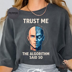 May include: Dark gray t-shirt with the text "TRUST ME" above a graphic of a human-robot hybrid face. The text "THE ALGORITHM SAID SO" is below the graphic. The shirt has a relaxed fit.