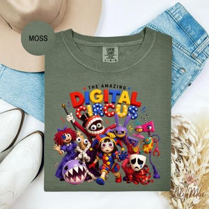 K&ouml;nnte beinhalten: Moosgr&uuml;nes Comfort Colors T-Shirt mit einer farbenfrohen Grafik von Cartoon-Zirkusfiguren. Der Text "THE AMAZING DIGITAL CIRCUS" ist &uuml;ber den Figuren zu sehen. Das Shirt liegt flach auf einer wei&szlig;en Oberfl&auml;che mit Hut, Stiefeln und Jeans.