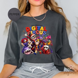 K&ouml;nnte beinhalten: Pfefferfarbenes Oversized-T-Shirt mit einem farbenfrohen Grafikdesign von Charakteren aus "The Amazing Digital Circus". Das Design enth&auml;lt den Text "THE AMAZING DIGITAL CIRCUS" in Rot, Blau und Gelb. Das Shirt hat eine lockere Passform.
