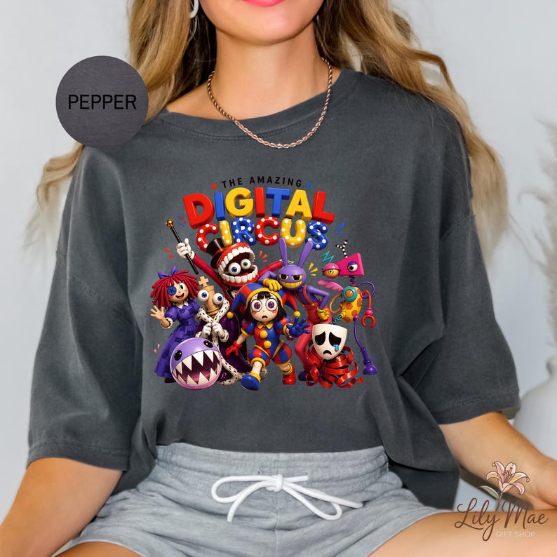 K&ouml;nnte beinhalten: Pfefferfarbenes Oversized-T-Shirt mit einem farbenfrohen Grafikdesign von Charakteren aus "The Amazing Digital Circus". Das Design enth&auml;lt den Text "THE AMAZING DIGITAL CIRCUS" in Rot, Blau und Gelb. Das Shirt hat eine lockere Passform.