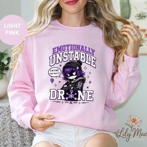 May include: Light pink sweatshirt with the text "Emotionally Unstable Drone" and a cartoon character graphic. The graphic includes the phrase "I'm fine (not really)". The sweatshirt is a crew neck style.