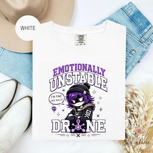 May include: White crew neck t-shirt with the text "Emotionally Unstable Drone" in purple. The graphic features a cartoon character with purple hair and a black beanie. The shirt also has the text "I'm fine (not really)".