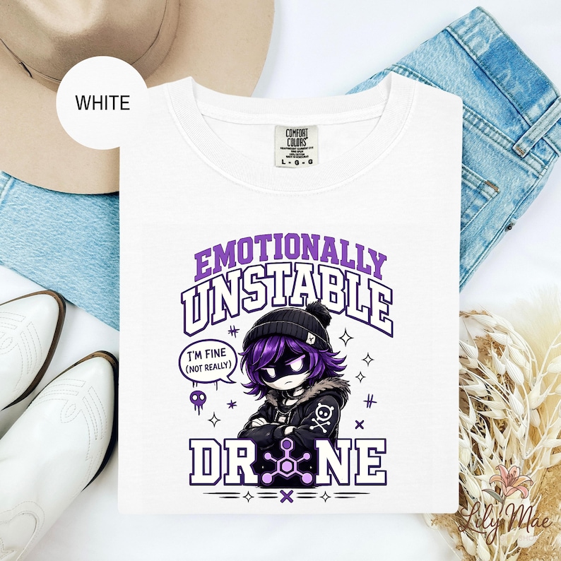 May include: White crew neck t-shirt with the text "Emotionally Unstable Drone" in purple. The graphic features a cartoon character with purple hair and a black beanie. The shirt also has the text "I'm fine (not really)".