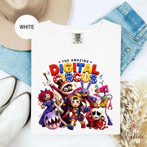 K&ouml;nnte beinhalten: Wei&szlig;es T-Shirt mit Rundhalsausschnitt und einem farbenfrohen Grafikdesign von Zeichentrickfiguren und dem Text "THE AMAZING DIGITAL CIRCUS". Das Design zeigt skurrile Figuren in leuchtenden Farben, ideal f&uuml;r Fans von Zeichentrickfilmen.