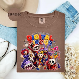 K&ouml;nnte beinhalten: Espressofarbenes T-Shirt mit einer farbenfrohen Grafik von Charakteren aus "The Amazing Digital Circus". Das Design enth&auml;lt den Text "THE AMAZING DIGITAL CIRCUS" in einer lebendigen, verspielten Schriftart. Das Shirt ist von der Marke Comfort Colors.