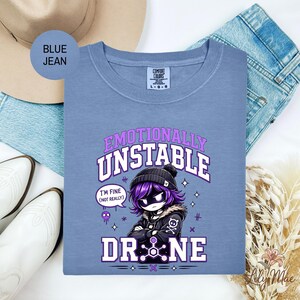 May include: A blue Comfort Colors t-shirt with the text "Emotionally Unstable" and a cartoon character. The character has purple hair and is wearing a black beanie and jacket. The text "I'm fine (not really)" is in a speech bubble. The word "DRONE" is also visible.