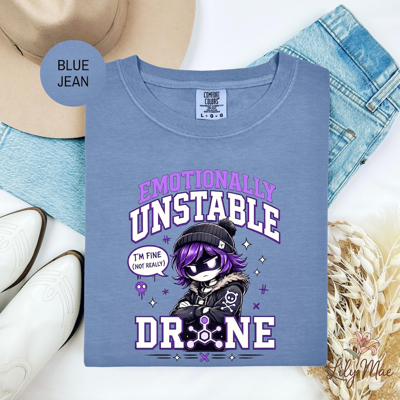 May include: A blue Comfort Colors t-shirt with the text "Emotionally Unstable" and a cartoon character. The character has purple hair and is wearing a black beanie and jacket. The text "I'm fine (not really)" is in a speech bubble. The word "DRONE" is also visible.