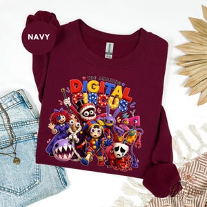 K&ouml;nnte beinhalten: Ein weinrotes Sweatshirt mit einer farbenfrohen Grafik von Charakteren aus "The Amazing Digital Circus". Das Design enth&auml;lt den Text "DIGITAL CIRCUS" in leuchtenden Farben sowie verschiedene Cartoon-Figuren. Das Sweatshirt ist aus weichem Material.