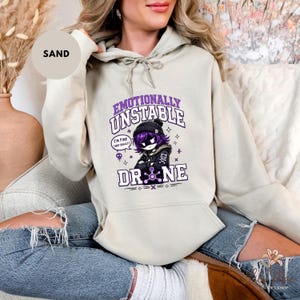 May include: Sand-colored hoodie featuring a graphic of an anime-style character with the text "Emotionally Unstable" and "Drone." The character has purple hair and is wearing a black beanie and jacket. The hoodie has a drawstring and a front pocket.