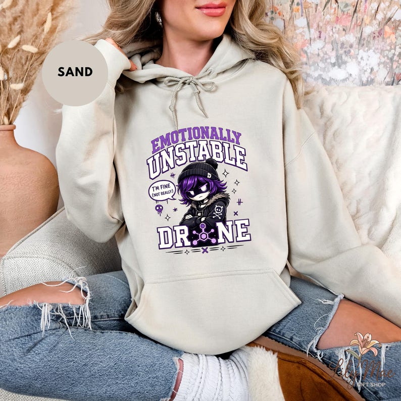 May include: Sand-colored hoodie featuring a graphic of an anime-style character with the text "Emotionally Unstable" and "Drone." The character has purple hair and is wearing a black beanie and jacket. The hoodie has a drawstring and a front pocket.