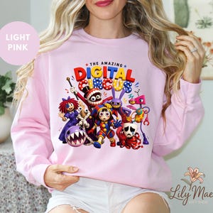 K&ouml;nnte beinhalten: Hellrosa Sweatshirt mit einem farbenfrohen Grafikdesign von Charakteren aus "The Amazing Digital Circus". Das Design zeigt Cartoonfiguren in leuchtenden Farben und den Text "The Amazing Digital Circus". Das Sweatshirt hat einen Rundhalsausschnitt.