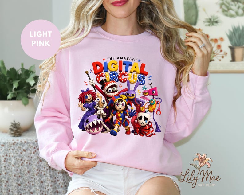 K&ouml;nnte beinhalten: Hellrosa Sweatshirt mit einem farbenfrohen Grafikdesign von Charakteren aus "The Amazing Digital Circus". Das Design zeigt Cartoonfiguren in leuchtenden Farben und den Text "The Amazing Digital Circus". Das Sweatshirt hat einen Rundhalsausschnitt.