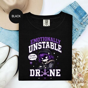 May include: Black crew neck t-shirt with a graphic design. The design features the words "Emotionally Unstable" in purple and white, along with a cartoon character and the word "DRONE". The character has purple hair and is wearing a black beanie.