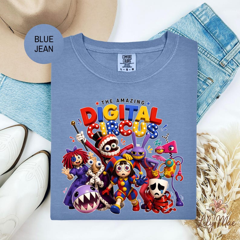 K&ouml;nnte beinhalten: Ein hellblaues Comfort Colors T-Shirt mit einer farbenfrohen Grafik von "The Amazing Digital Circus". Das Design zeigt verschiedene Cartoon-Charaktere und den Text "THE AMAZING DIGITAL CIRCUS" in leuchtenden Farben. Das T-Shirt ist von einem Hut, Jeans und wei&szlig;en Stiefeln umgeben.