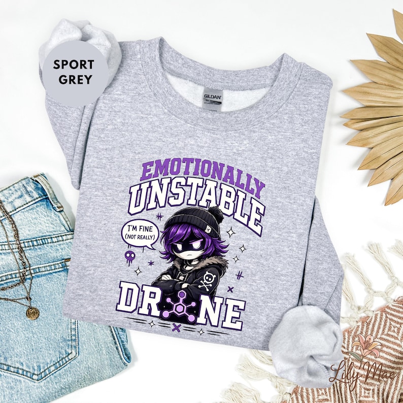 May include: Sport grey sweatshirt with the text "Emotionally Unstable Drone" and a graphic of a character with purple hair and a black beanie. The character has a black jacket and the words "I'm fine (not really)" in a speech bubble.