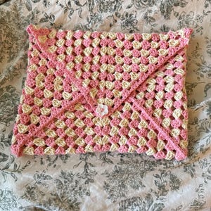 May include: A handmade crochet envelope-style clutch in a pink and cream granny square pattern. The clutch is secured with a pink flap and a white flower-shaped button. The item is made of yarn and is suitable for carrying small items.