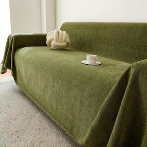 May include: A green sofa covered with a matching throw blanket. A white knot-shaped pillow and a white teacup and saucer sit on the sofa. The sofa is in a well-lit room with a neutral-colored carpet.