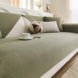 May include: A sage green textured sofa cover draped over a white couch, featuring a matching sage green throw pillow, a rectangular light gray cushion, a round decorative ball-shaped pillow, and a white ceramic mug on a small matching saucer.