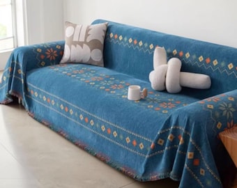 May include: A blue sofa cover featuring a repeating pattern of orange and yellow diamonds, zig-zag lines, and floral motifs. The cover is draped over a sofa, accompanied by a beige throw pillow with white shapes, a white knot pillow, and a white mug.