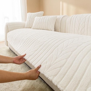 May include: A cream-colored sofa cover featuring a textured, raised leaf-like pattern. The cover is draped over a sofa, with matching rectangular and square throw pillows placed on the back cushions, all showcasing the same consistent design.
