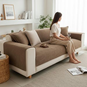 May include: A three-seater sofa featuring a cream-coloured frame and a textured brown fabric cover draped over the seat and backrest. The sofa includes two matching brown throw pillows and one cream-coloured throw pillow, providing a neutral aesthetic.