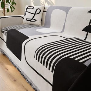 Puede incluir: Una manta para sofá y un cojín cuadrado a juego con un patrón geométrico en negro, blanco y gris. El diseño textil incluye bloques negros sólidos, rayas negras paralelas finas y formas redondeadas en gris suave sobre una base blanca.