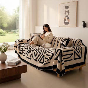 Puede incluir: Una manta para sofá de color crema con un llamativo patrón geométrico negro de triángulos y líneas. El tejido rectangular tiene un borde negro grueso y está colocado sobre un sofá de madera, aportando un elemento visual de alto contraste.