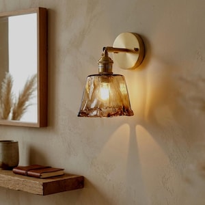 Op de afbeelding: Een wandlamp met een messing afwerking en een amberkleurige glazen kap met textuur. Het licht is aan en geeft een warme gloed. De armatuur is gemonteerd op een beige muur, met een houten plank en spiegel op de achtergrond.