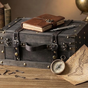 May include: A dark wooden chest with metal corner reinforcements and leather straps, topped with a brown leather-bound journal and a silver pen. Beside the chest lies a brass compass, a vintage-style map, and several small metal keys.