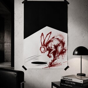 May include: A framed art print featuring a red rabbit leaping over a black hole. The artwork is set against a white and black geometric background, attached to a textured wall with black tape. A black table and lamp are in the foreground.