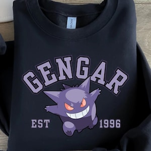 Könnte beinhalten: Schwarzes Sweatshirt mit einem lila Gengar-Motiv. Das Design beinhaltet das Wort "GENGAR" in einer College-Schriftart und "EST 1996" darunter. Das Motiv zeigt einen lächelnden Gengar-Charakter mit roten Augen.
