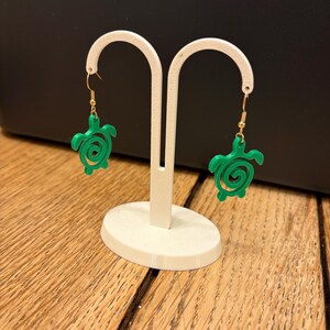 Puede incluir: Pendientes verdes con forma de tortuga y ganchos dorados. Se exhiben en un expositor de joyas blanco sobre un fondo oscuro. El diseño de la tortuga presenta un patrón en espiral en el caparazón.