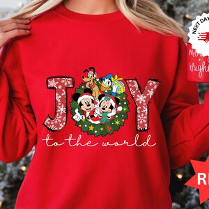 Puede incluir: Una sudadera roja con un diseño navideño. Presenta a Mickey Mouse, Minnie Mouse, el Pato Donald y Pluto. La palabra "JOY" está decorada con copos de nieve, y la frase "to the world" está escrita debajo. Una taza roja se sostiene en la mano.