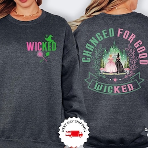 May include: Dark grey heather sweatshirt with the word "WICKED" in pink and green on the front, along with a witch hat and lollipop graphic. The back features the phrase "CHANGED FOR GOOD" and a graphic of two women in front of a castle.