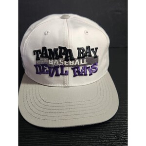 May include: A white and gray baseball cap with the text "TAMPA BAY BASEBALL DEVIL RAYS" embroidered on the front. The cap has a gray brim and a white crown. The text is in black and purple.