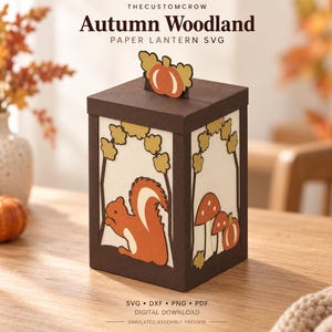 May include: A square paper lantern featuring a dark brown frame and cream-coloured panels with cut-out designs of a squirrel, mushrooms, and a pumpkin. The top lid is adorned with a small pumpkin and leaf cutout. Text reads THECUSTOMCROW Autumn Woodland PAPER LANTERN SVG.