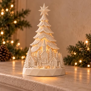 May include: A cream-colored, illuminated Christmas tree figurine featuring a star topper and a detailed winter village scene at the base, including a church, small houses, and evergreen trees, all set on a faceted circular pedestal.