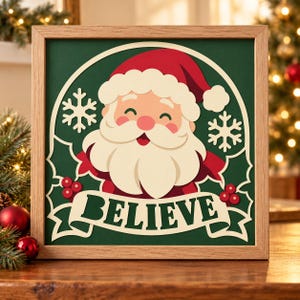 May include: A square wooden framed wall art piece featuring a layered paper cutout of Santa Claus on a dark green background. Santa wears a red hat and has a white beard. The design includes white snowflakes, holly berries, and a banner with the text BELIEVE.